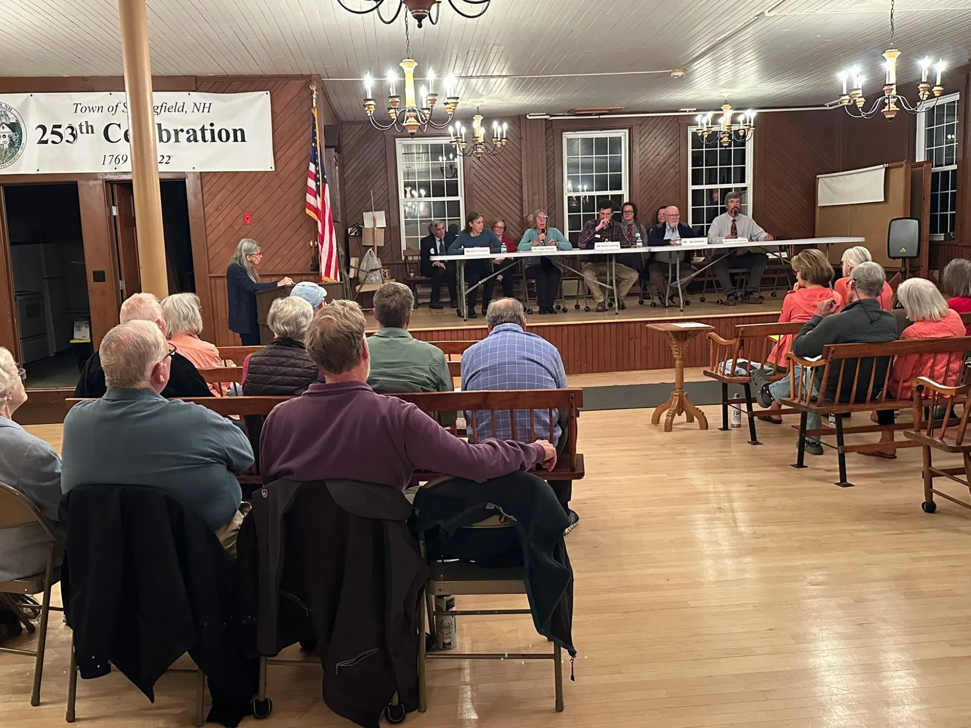 A packed Springfield town hall, residents filling the pews while candidates sit at a long table on the stage