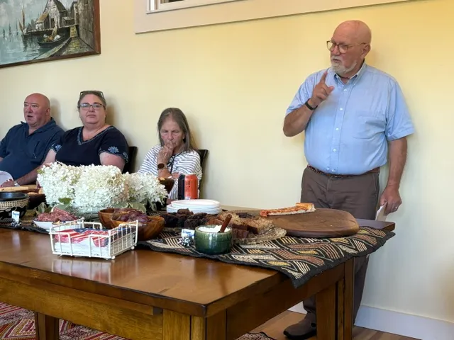 Jim Bednar gesturing while speaking, standing behind a table set with food, guests listening