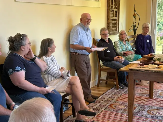 Jim Bednar standing with notes in hand, speaking to a seated group in a sunlit room